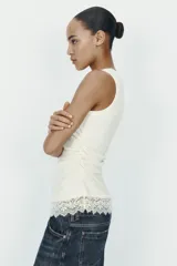 Camiseta ajustada (slim fit) color crema, con cuello redondo y mangas sisa. Presenta frunces laterales y un bajo asimétrico con detalle de encaje a tono.