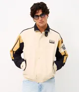 Campera de sarga de algodón marca Blue Steel, con diseño de bloques de color en tonos beige, negro y mostaza. Presenta cuello alto, cierre frontal con botones a presión y solapa, bolsillos laterales y detalles de parches bordados con temática outdoor en el pecho y la manga.