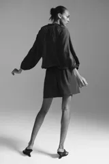 Vestido túnica corto color marrón en mezcla de lyocell y viscosa con leve brillo. Modelo oversized con cuello redondo con ribete trenzado grueso, escote en V con tiras finas, mangas globo largas con abertura en los hombros, elástico en los puños y cintura fruncida con vuelo en la falda.