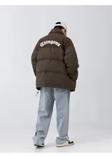 Campera puffer marrón con paneles horizontales acolchados. La parte superior de la espalda es de pana marrón con el texto 'Chingona' bordado en blanco. Posee cordón ajustable en el ruedo y se lleva superpuesta a una sudadera con capucha gris.