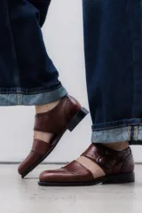 Zapato tipo monk strap con aberturas laterales, confeccionado en cuero vacuno metalizado color bronce. Presenta detalles de perforaciones estilo brogue en la puntera y el talón, y doble hebilla metálica en el empeine.