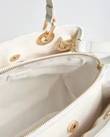 Cartera tipo bolso de mano XL color blanco de símil cuero con diseño matelassé romboidal, tachas metálicas doradas, doble asa corta y correa bandolera regulable y desmontable. Incluye herrajes dorados y logo metálico de la marca en el frente.
