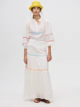 Falda larga blanca confeccionada en broderie de algodón con diseño de paneles escalonados, detalles de galones con transparencias en color azul y naranja, y pretina elástica con cordón ajustable.