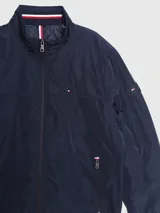 Campera bomber Tommy Hilfiger azul marino con cuello alto, cierre frontal con cremallera, dos bolsillos laterales con cremallera, puños y bajo elásticos. Tiene el logo de Tommy Hilfiger bordado en el pecho y un parche con el logo en la manga izquierda. El interior del cuello tiene detalles en rojo, blanco y azul.