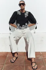 Blusa corta negra con mangas abullonadas y bordados florales en contraste de color blanco. Presenta cuello redondo con puntilla, mangas ajustables con cordones y borlas, y cierre en la espalda con abertura y botón oculto.