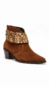 Bota corta de gamuza color marrón, con taco medio ancho y punta afinada. Cuenta con un aplique desmontable de apliques metalizados dorados alrededor del tobillo.