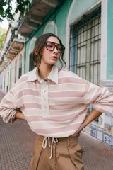 Remera tipo polo de tejido de punto a rayas horizontales en tonos rosados y beige. Presenta cuello camisero de lino, cierre frontal con botones de nácar y silueta holgada.