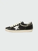 Zapatillas Golden Goose modelo Super Star, confeccionadas en terciopelo negro con acabado desgastado. Presentan la icónica estrella lateral en cuero plateado, cordones gruesos en color crema y suela de goma con efecto envejecido.
