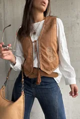 Chaleco de gamuza color camel con bordados decorativos en hilo claro y cierre frontal mediante lazos con borlas. Presenta un diseño de inspiración boho con terminaciones delicadas.