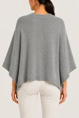 Poncho de punto color gris, con diseño de corte asimétrico, cuello redondo y cierre frontal con botones metálicos. Presenta terminaciones en tejido acanalado en el cuello, los bordes laterales y el ruedo.