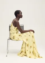 Vestido largo de tirantes finos con cuello de pico, confeccionado en viscosa con estampado floral difuminado en tonos amarillos y marrones. Presenta un diseño recto con detalle de cadena metálica a la altura de la cadera y cierre de lazo en la espalda.