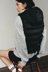 Chaleco acolchado de color negro con cuello subido, capucha ajustable y cierre frontal con cremallera termosellada. Presenta un diseño con paneles laterales en tejido liso y bolsillos laterales.