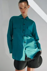 Camisa de corte fluido con cuello solapa y manga larga. Presenta cierre frontal con botones y dos bolsillos de plastrón con solapa en el pecho. El diseño cuenta con bajo redondeado y costuras verticales decorativas.