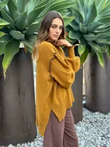 Sweater de lana color mostaza, con cuello redondo y mangas largas abullonadas. Presenta detalles de costuras expuestas en color marrón oscuro en las mangas y el cuello. El dobladillo es asimétrico con aberturas laterales.