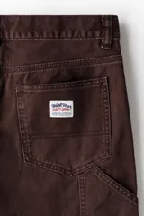 Pantalón estilo carpenter de sarga de algodón, color marrón oscuro, con corte recto y costuras reforzadas en las rodillas. Presenta bolsillos laterales, bolsillo monedero, bolsillos traseros, trabilla lateral y bolsillo cargo con doble compartimento.
