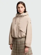 Campera corta con capucha, color beige, de corte holgado y diseño minimalista. Presenta cierre frontal oculto, bolsillos laterales con solapa y botones a presión, mangas largas con puños ajustables y cordones decorativos en la capucha. Incluye el logo de la marca en la manga izquierda.