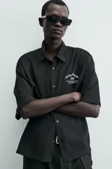 Camisa de corte relajado confeccionada en tejido fluido con mezcla de viscosa, color negro. Presenta cuello solapa, manga corta, cierre frontal con botones y un bordado a contraste en el pecho.