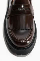 Mocasines tipo penny de color marrón oscuro, confeccionados en material sintético con acabado brillante. Presentan flecos decorativos en el empeine, una banda transversal con detalle metálico y una suela gruesa dentada de color negro.