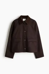 Campera de sarga color marrón oscuro, con cuello camisero de pana, cierre frontal oculto por solapa con botones a presión y bolsillos frontales grandes con solapa.