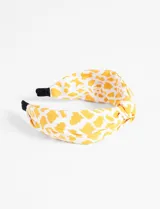 Vincha rígida de tela con nudo superior y estampado de manchas color mostaza sobre fondo blanco.