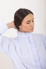 Camisa de manga larga a rayas celestes y blancas, con cuello alto mao y volados frontales a lo largo de la botonadura.