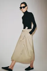 Falda midi color beige confeccionada en hilatura con mezcla de algodón. Presenta tiro medio, cintura con trabillas, bolsillos laterales ocultos en costura y cinturón del mismo tejido. Su diseño incluye un bajo acabado en línea evasé y cierre lateral con cremallera.