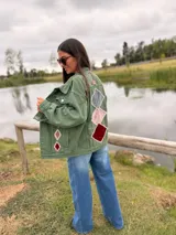 Campera de jean color verde oliva, con diseño de figuras geométricas en terciopelo aplicadas en la espalda y mangas, bordadas a mano con costuras decorativas. Presenta un corte holgado y etiqueta de cuero en la parte superior de la espalda.