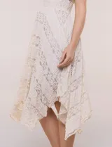 Vestido midi blanco de estilo romántico confeccionado en poliéster con elastano, con detalles de encaje floral. Cuenta con escote cruzado en V, mangas cortas acampanadas, cintura elastizada y falda evasé con terminación asimétrica en picos.