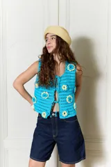 Chaleco tejido en crochet color celeste, de calce relajado y sin mangas, con diseño abierto en el frente y cierre mediante lazos. Presenta aplicaciones de flores en relieve de color amarillo y blanco.