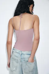 Body de tejido elástico color rosado con escote redondo y tirantes finos. Presenta un detalle de encaje a tono en la parte superior del escote que se ajusta mediante una lazada al cuello. Cuenta con cierre en el bajo mediante botones a presión.