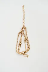 Brazalete multicapa de color dorado compuesto por cadenas de eslabones tipo caja y cadenas de esferas pequeñas, con un dije en forma de cubo en el extremo.