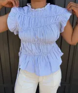 Blusa sin mangas a rayas finas en celeste y blanco, con cuello redondo alto y volados en los hombros. Presenta frunces en el frente, lazos laterales regulables y bajo con ligero vuelo.