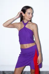 Falda corta violeta de diseño cruzado estilo pareo, con cierre lateral invisible y un lazo rojo largo que cae desde el lateral.