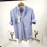Camisa de vestir a rayas verticales azules y blancas, de corte clásico, con cuello italiano y mangas largas con puños abotonados. Presenta logo bordado en el pecho. Marca Duxon.