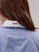 Camisa de manga larga en color azul con textura tipo denim, con cuello y puños en contraste de color blanco. Presenta costuras decorativas horizontales y verticales en el frente, cierre frontal con botones y corte recto.