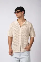 Camisa de punto calado en color beige, de manga corta y corte relajado. Presenta cuello tipo bowling, cierre frontal con botones al tono y una estructura liviana y respirable confeccionada en algodón.