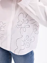 Camisa beige de manga larga con cuello camisero, cierre frontal con botones y diseño de pespuntes y bordados en relieve con formas florales en la parte inferior y lateral.