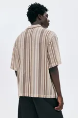 Camisa de manga corta con cuello inglés y botones frontales, confeccionada en tejido de punto pointelle con patrón de rayas verticales en tonos tierra y beige.