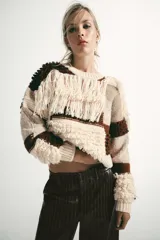 Jersey de punto con cuello redondo y manga larga, caracterizado por un diseño de parches en tonos tierra y crema. Presenta una textura variada con detalles de flecos y tejido en relieve.