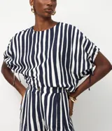 Blusa de viscosa con estampado de rayas verticales blancas y azul marino. Tiene cuello redondo, mangas cortas con detalle de lazo ajustable en los laterales y cierre de botón en la espalda.