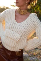 Buzo tejido a mano en lana merino color crudo, con capucha, escote en V y bolsillo canguro frontal.