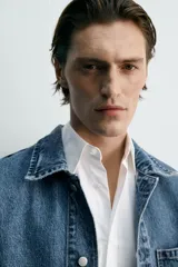 Cazadora de denim de algodón, color azul medio lavado, con cuello solapa, manga larga y cierre frontal de botonadura. Presenta cuatro bolsillos de plastrón con tapa y botón en el frente (dos en el pecho y dos a la altura de la cadera).