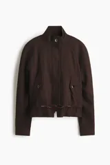 Campera corta oversize de color marrón, confeccionada en tejido de lino. Presenta cuello elevado, cierre frontal oculto con solapa y botones visibles, mangas raglán con puños abotonados, bolsillos delanteros con solapa y cordón ajustable en el bajo.
