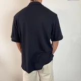 Camisa de manga corta tejida en color negro, con cuello tipo camp y cierre frontal de botones blancos. Presenta un calce holgado y un pequeño logo bordado en el pecho.