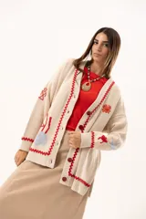 Saco tipo cardigan oversize de punto color beige, con diseño temático marino que incluye bordados de corales, peces, estrellas de mar y tortugas. Presenta detalles de galón picot en color rojo en los bordes y puños, cierre frontal con botones al tono y corte holgado.