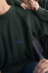 Buzo de punto acanalado color verde oscuro, de manga larga con diseño raglán, cuello redondo y un pequeño parche bordado en el pecho. Presenta detalles de rayas en los puños y el ruedo.