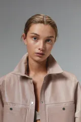 Camisa de cuero ovino color rosa pálido, de corte recto y estilo minimalista. Tiene mangas cortas, cuello clásico, cierre con botones a presión plateados y bolsillos delanteros con solapa.