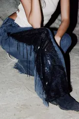 Jeans de corte wide leg en denim negro lavado, con inserción lateral de encaje negro adornado con flecos y lentejuelas.
