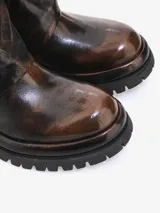 Bota corta de cuero bovino con efecto desgastado en tonos negro y gris. Presenta un diseño robusto con caña arrugada y una cremallera diagonal decorativa con tirador dorado. La suela es gruesa y dentada.