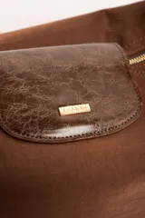 Cartera tipo shopper color marrón, con solapa frontal decorativa en tono más oscuro y tiras con nudos. Incluye herrajes dorados y un charm colgante con cuentas de madera.